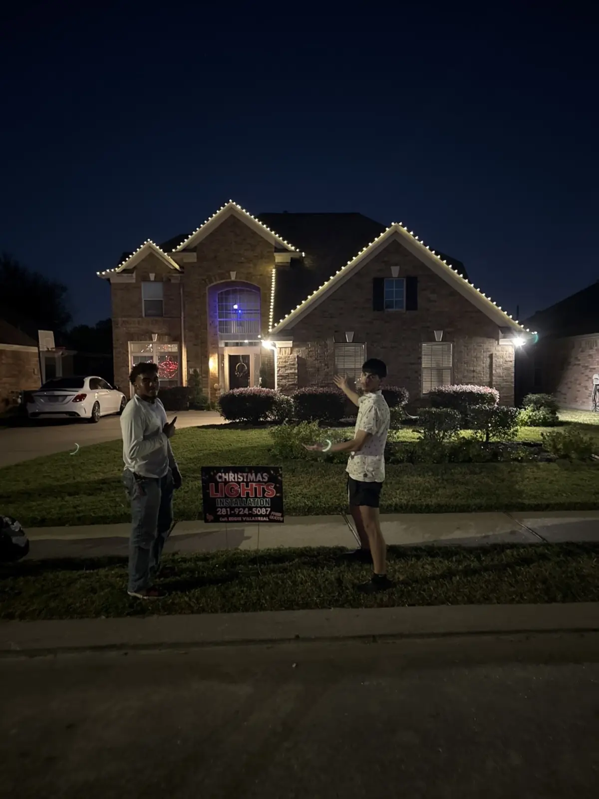 Two-story house with warm white Christmas lights along the roof, crew standing out front